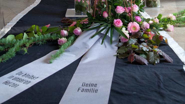 Blumenarrangement mit rosa Rosen und eine Trauerschleife vor einem Kreuz in einer Kapelle.