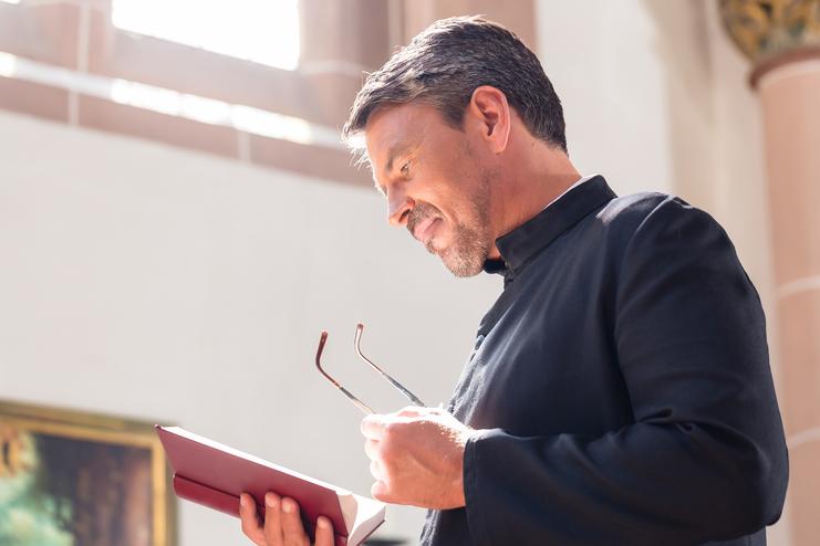 Ein Mann in schwarzer Robe liest aufmerksam in einem Buch, während er eine Brille in der Hand hält.