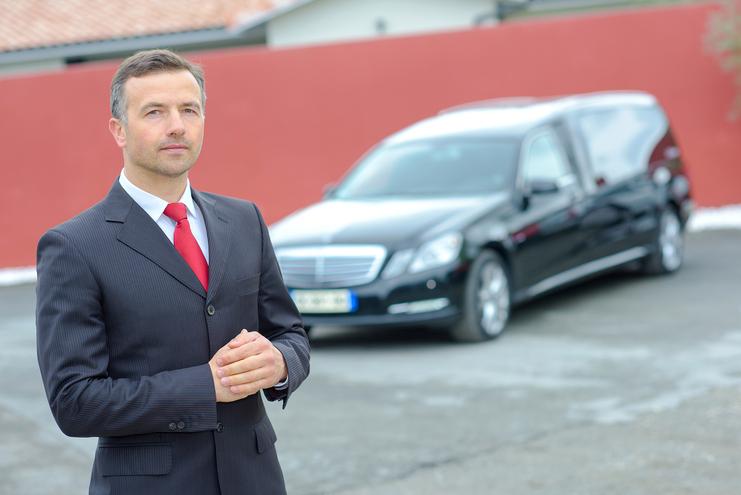 Mann im Anzug mit roter Krawatte steht vor einem schwarzen Leichenwagen auf einem befestigten Platz.
