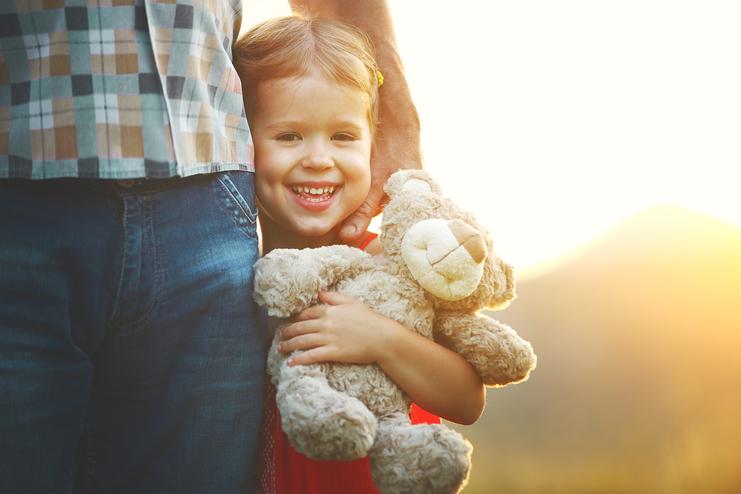 Lächelndes Kind mit einem Plüschteddy steht neben einem Erwachsenen, vor einer sonnigen, verschwommenen Landschaft.
